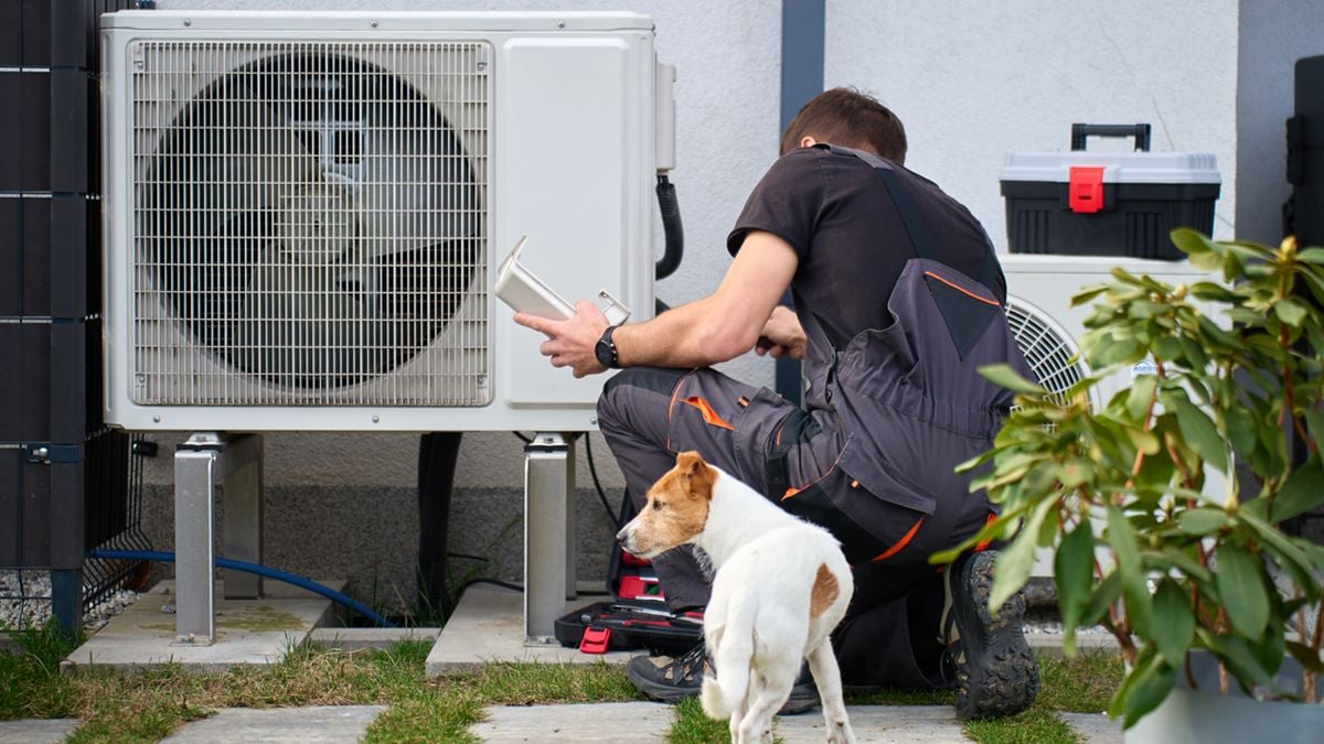 Pompe &agrave; chaleur et assurance habitation : un technicien contr&ocirc;le l'unit&eacute; ext&eacute;rieure d'une PAC devant une maison, sous l'&oelig;il curieux d'un petit chien