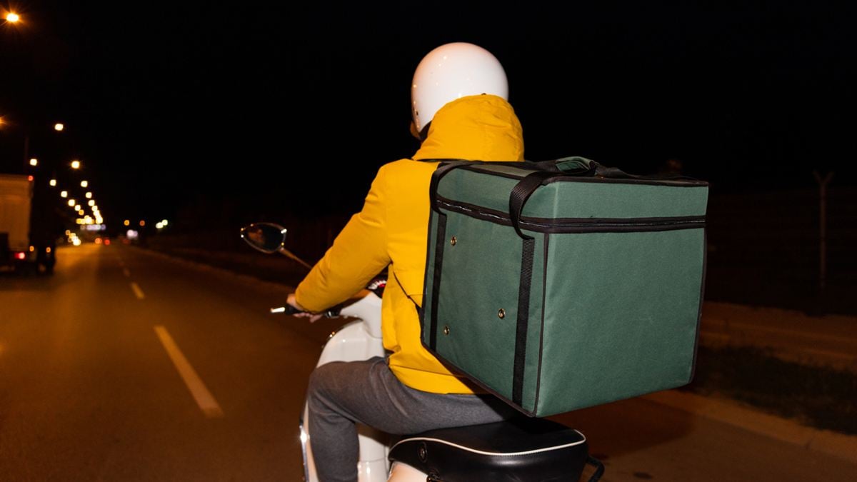 Assurance deux-roues pour livreur : de nuit, un livreur en veste jaune et casque blanc roule en scooter, sac isotherme vert sur le dos