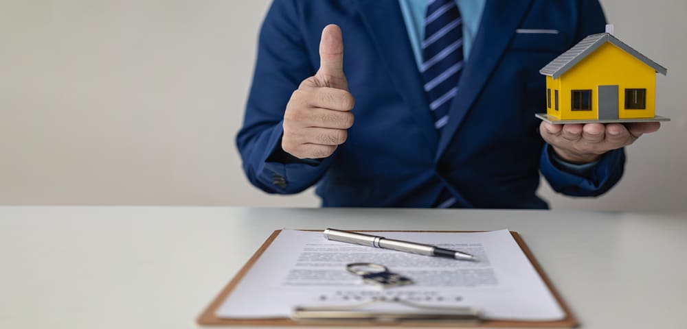 Un homme en costume, représentant un auto-entrepreneur, tient une maison miniature et fait un pouce levé devant un contrat de crédit immobilier signé, symbolisant l’obtention d’un prêt immobilier.