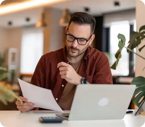 Un homme portant des lunettes consulte des documents devant un ordinateur portable dans un bureau lumineux